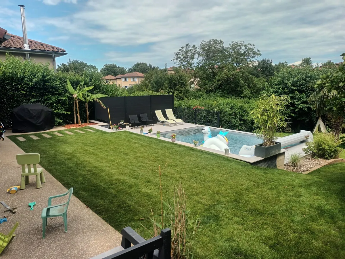 Piscine rectangulaire bordée d'une pelouse, terrasse et transats, jardin arboré en arrière-plan