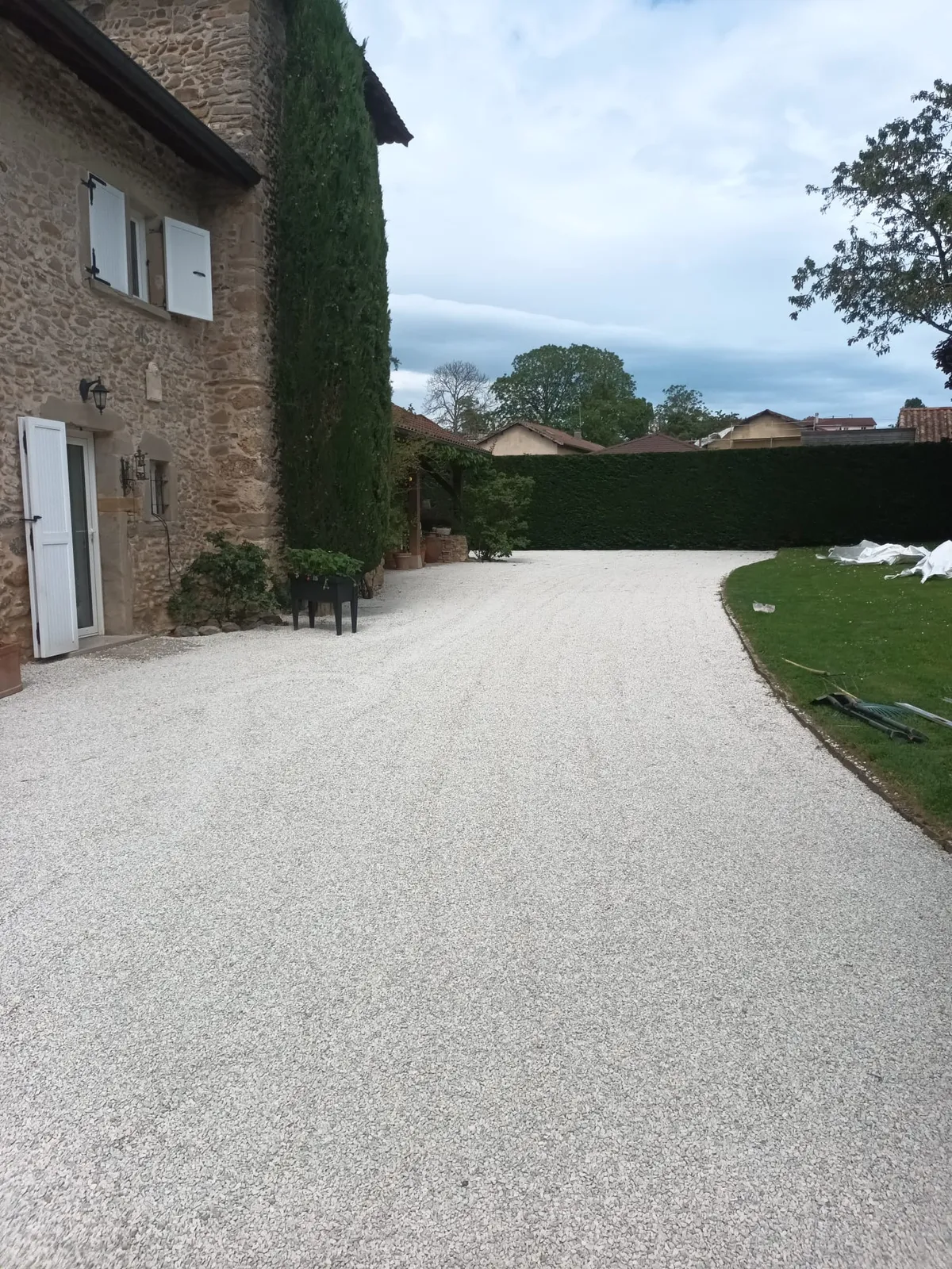 Allée en gravillon lavé clair devant une maison en pierre, haie taillée en fond